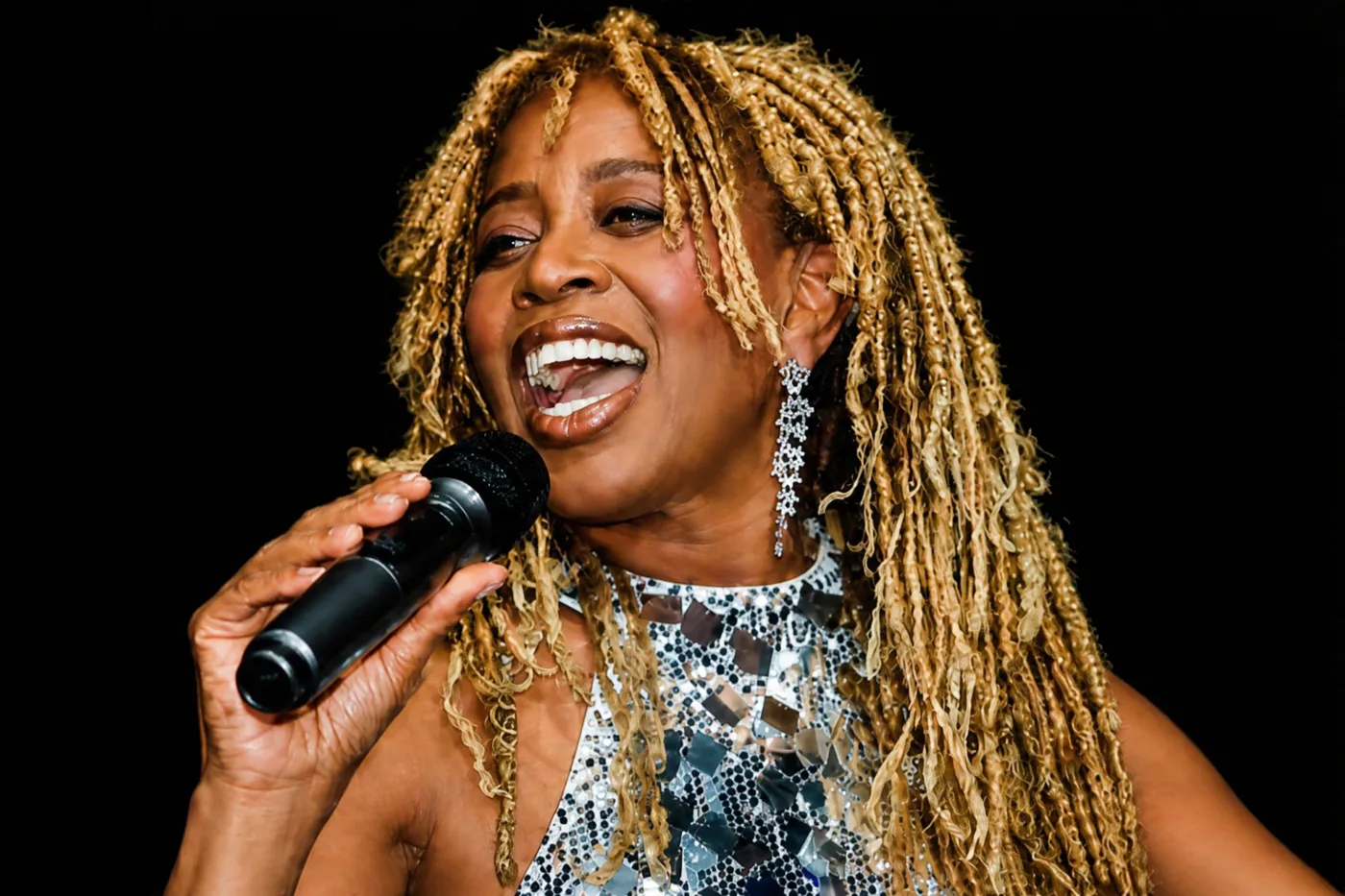 Close-up of JB Rose singing into a microphone, wearing long drop earrings and a mirrored halter-neck dress
