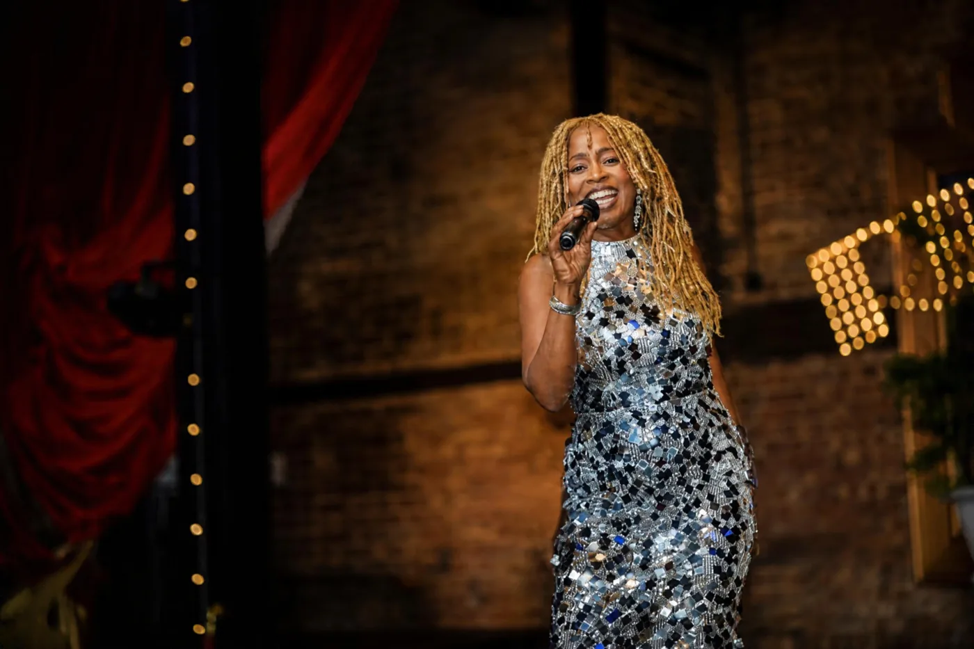 JB Rose singing on a stage with a brick wall, red curtain and strings of lights behind her
